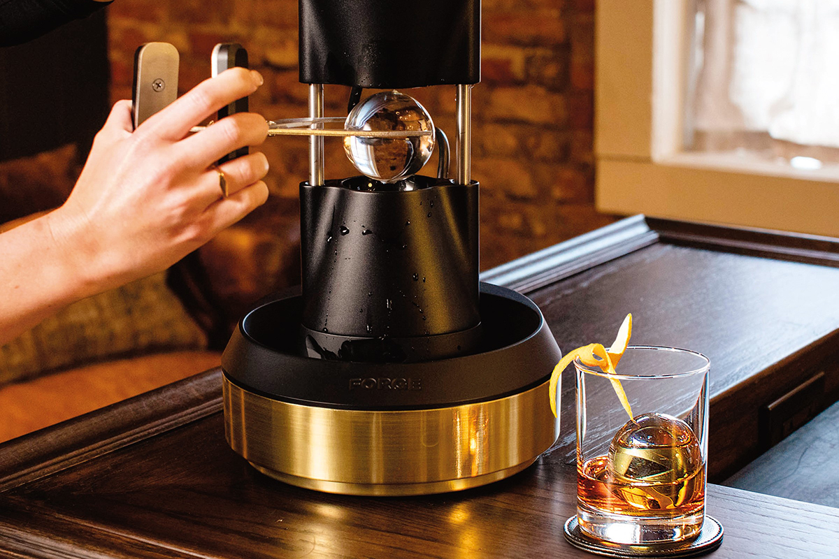 Person removing an ice sphere from a heated ice press made from die-cast anodized aluminum. There is a cocktail with citrus garnish and ice sphere next to the press.