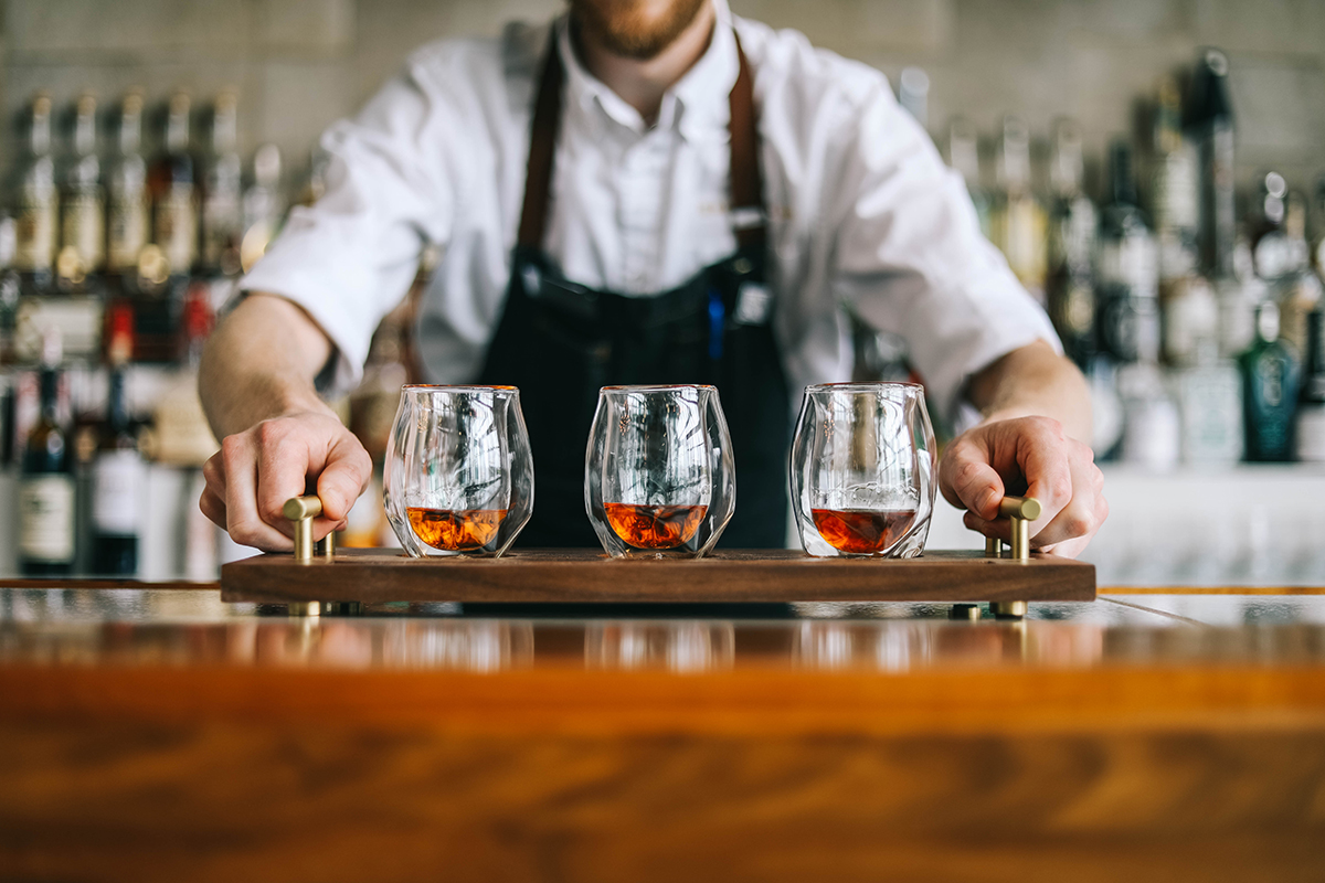 A Bardstown Bourbon Company bartender wearing a white shirt with apron serving samples of whiskeys on a tray. He is standing at bar with bottles in the background.