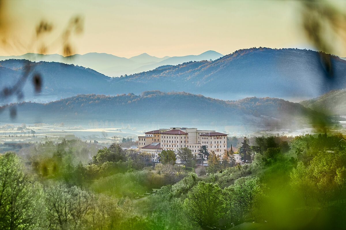 An exterior view of Palazzo Fiuggi, a wellness retreat located in the Italian countryside. The building is a historic stone palace with beige and brown hues, and it is surrounded by green trees and bushes. 