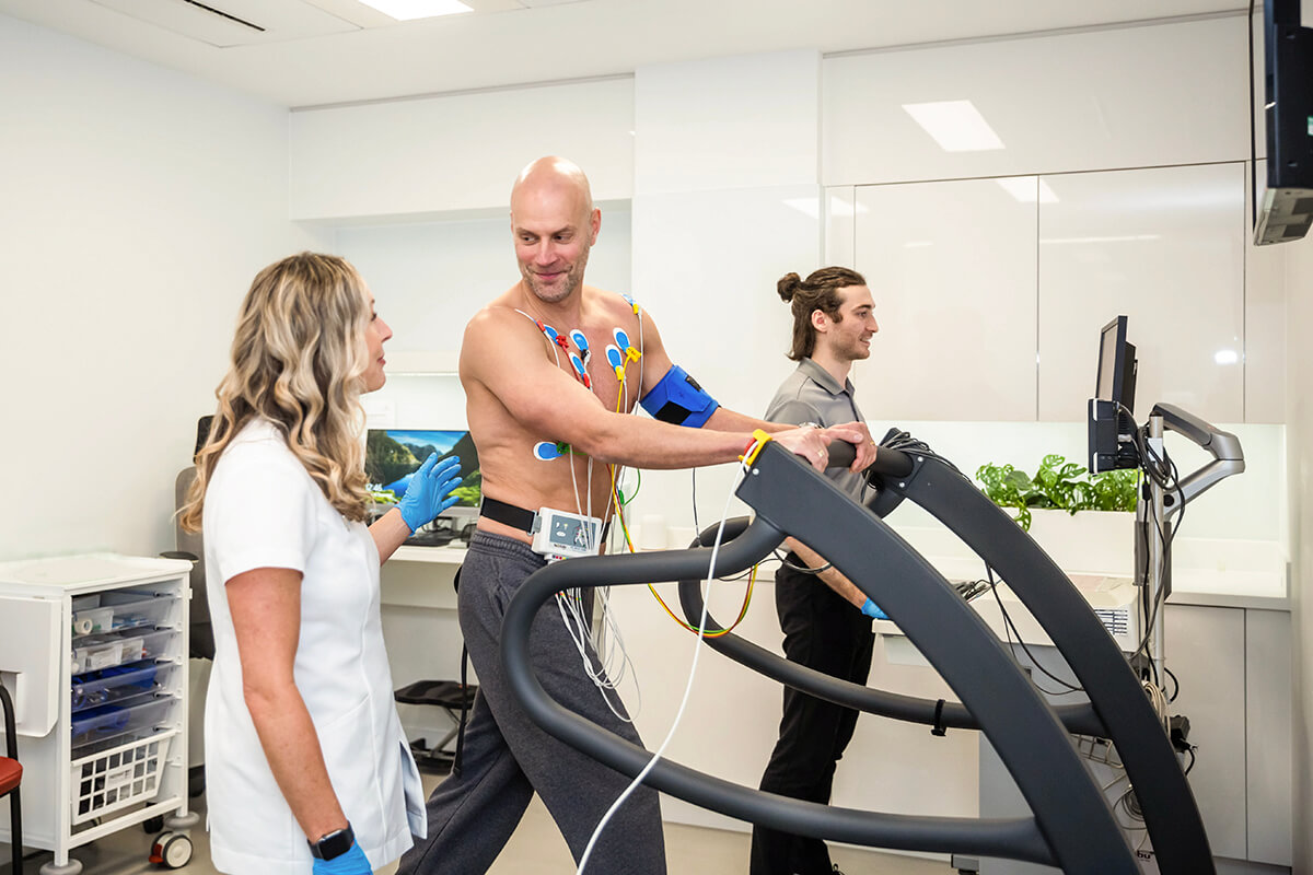 A person undergoing an exercise stress test at a medical facility while attached to various monitoring devices. The person is walking on a treadmill while a medical professional monitors their heart rate, blood pressure, and other vital signs.