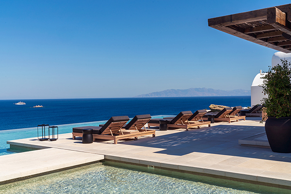 A beautiful infinity pool with a stunning view overlooking the sea on the island of Mykonos in Greece. The pool is surrounded by sun loungers. The blue sky and sea contrast with the white architecture of the surrounding buildings, creating a picturesque and serene scene. 