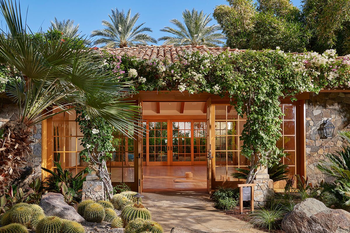 A spacious outdoor pavilion surrounded by green trees and vegetation. The pavilion features a wooden floor, wooden roof with a pointed tip, and wooden pillars supporting the structure. In the center of the pavilion, there are yoga mats placed on the wooden floor. The overall ambiance is serene and peaceful, making it an ideal setting for practicing yoga and meditation.