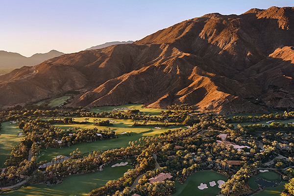 An aerial view of the Sensei Porcupine Creek Resort in Palm Springs, California. The resort is set amidst a sprawling desert landscape, surrounded by mountains and lush greenery. The image captures the beautiful architecture of the resort, with several buildings, a golf course, and a large swimming pool visible. 