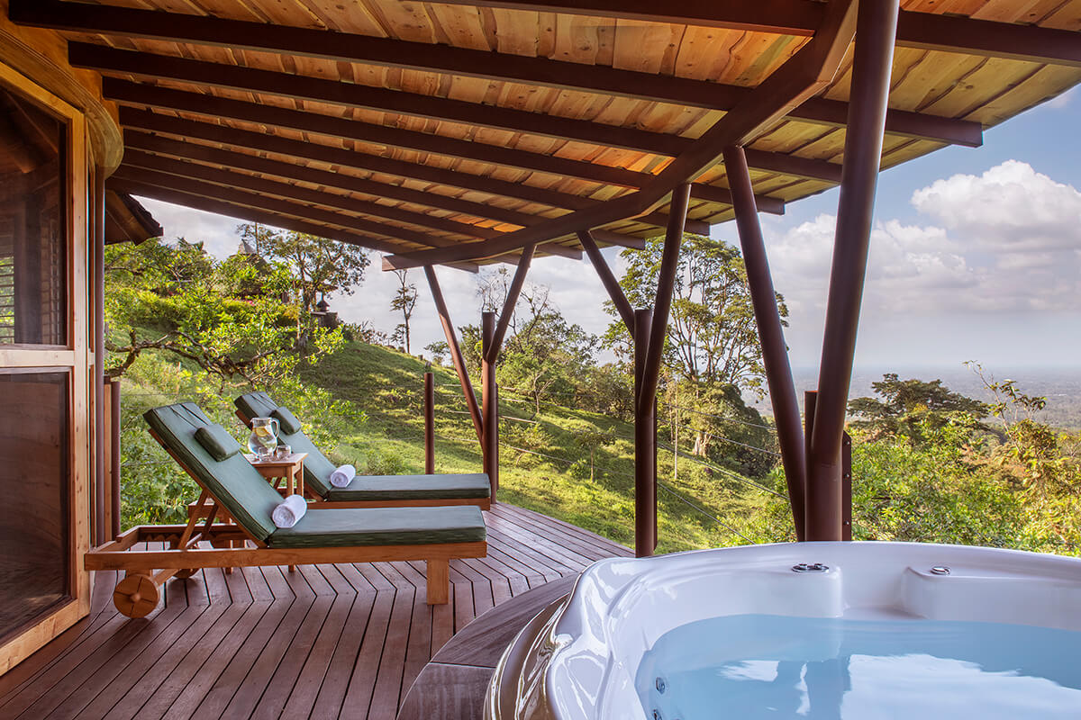 A luxurious outdoor spa area with a wooden deck surrounded by lush greenery and tropical plants. In the center of the deck is a large jacuzzi with bubbling water. The sky is blue with some fluffy white clouds, and the sun is shining, creating a warm and inviting atmosphere.